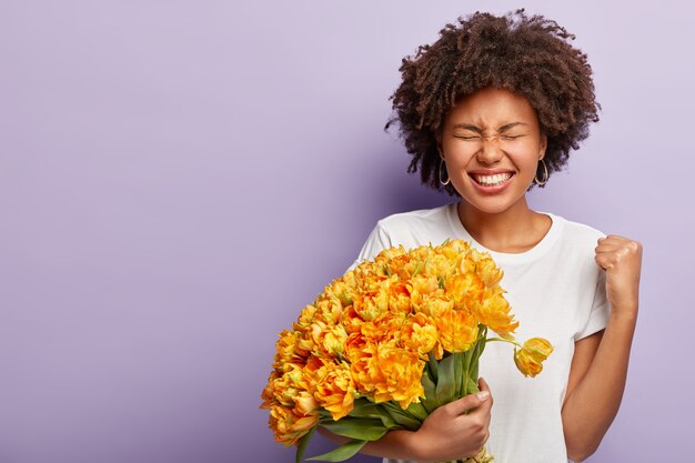 Retrato de dama triunfante con piel oscura, recibe un bonito ramo de flores de su amante, aprieta los puños, se ríe alegremente