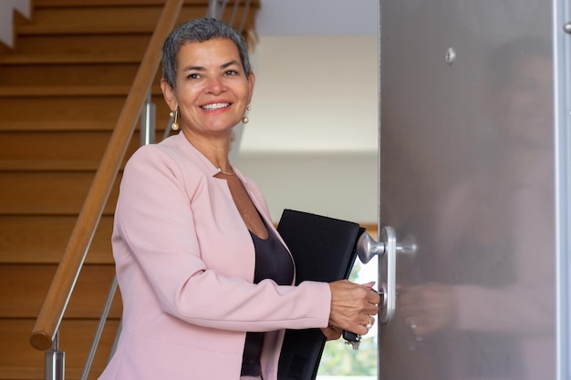 Retrato de dar la bienvenida a un agente inmobiliario en la entrada de la casa. Mujer sonriente con el pelo corto y gris en traje rosa preparándose para conocer a los clientes. Bienes raíces, negocios, concepto de trabajo.