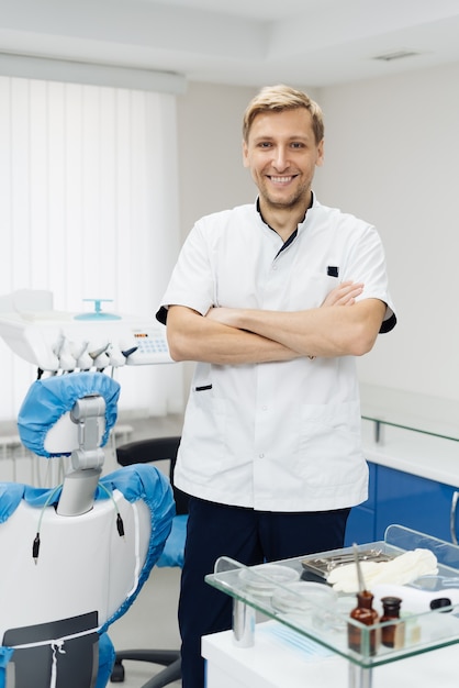 Retrato de un dentista masculino joven positivo en uniforme con las manos cruzadas en el consultorio dental