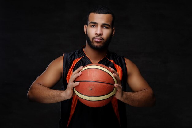 Retrato de un deportista afroamericano. Jugador de baloncesto en ropa deportiva con una pelota sobre un fondo oscuro.