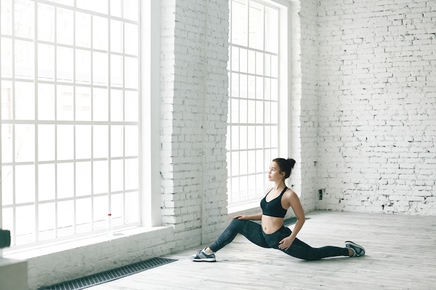 Foto gratuita retrato de deportista caucásica joven flexible fuerte en traje deportivo de moda haciendo pose de estiramiento, preparándose para divisiones frontales. atractiva chica en forma haciendo ejercicios para fortalecer la salud pélvica