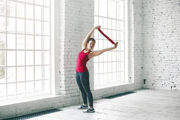 Retrato deportivo joven atractiva estudiante de yoga principiante en ropa deportiva con estilo haciendo asanas en la espaciosa habitación ayudándose a sí misma con la correa. Personas, deportes, fitness, yoga, pilates y estilo de vida activo