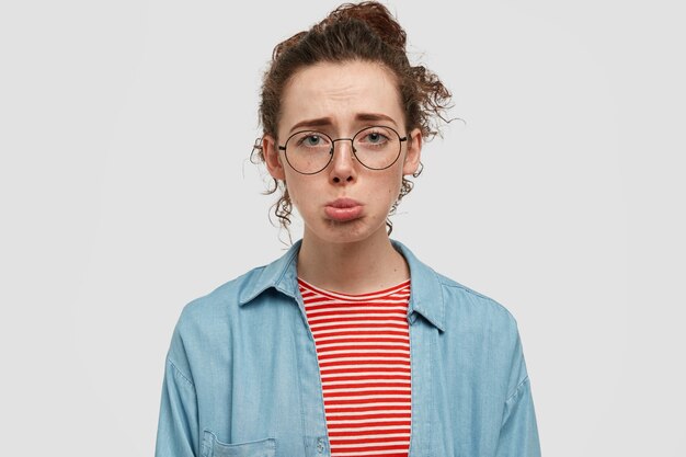 Retrato de descontento adolescente pecoso con gafas posando contra la pared blanca