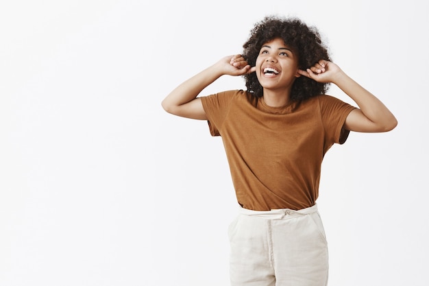 Retrato de despreocupada, indiferente y feliz joven afroamericana en camiseta marrón que cubre las orejas con los dedos índices mirando hacia arriba con alegría con una amplia sonrisa escuchando fuerte explosión