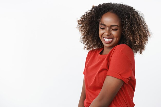 Retrato de despreocupada y tonta feliz hermosa y sincera joven afroamericana agitando la cabeza y saltando con los ojos cerrados y una amplia sonrisa alegre sobre la pared blanca