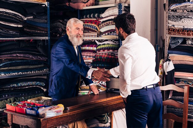 Retrato de un diseñador de moda masculino mayor sonriente que sacude las manos con el cliente en su tienda