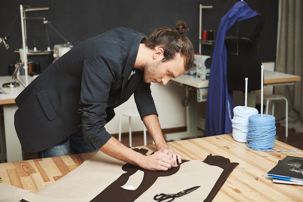 Retrato del diseñador de ropa masculina apuesto y masculino con un elegante peinado en traje negro cortando partes del futuro vestido de tela. El hombre se concentró en el trabajo.