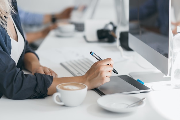 Retrato de diseñador web independiente tomando café en el lugar de trabajo y sosteniendo el lápiz. Señora ligeramente bronceada en camisa negra con tableta en su oficina, sentada frente a la computadora.