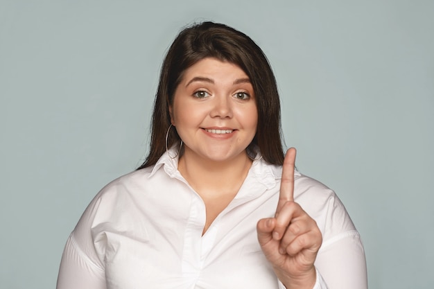 Retrato de divertida hermosa mujer joven de talla grande con camisa blanca mostrando el dedo índice como si tratara de llamar su atención, sonriendo ampliamente. Emociones, sentimientos y reacciones humanas