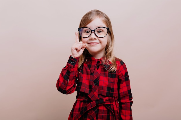 Retrato de divertida niña inteligente lleva gafas y camisa a cuadros levantó un dedo y sonríe en el frente