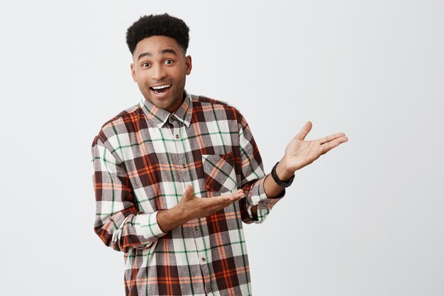 Retrato de divertido joven hermoso de piel oscura con peinado afro en camisa casual sonriendo, mostrando la pared blanca con las manos con expresión emocionada y feliz