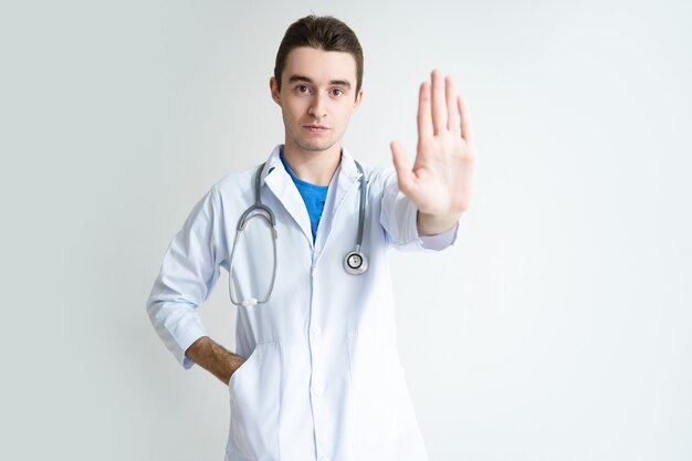 Retrato del doctor de sexo masculino joven serio que muestra el gesto de la parada
