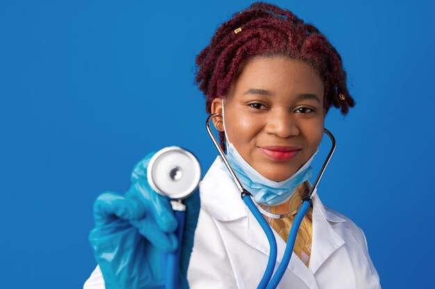 Retrato de doctora africana en bata de laboratorio con mascarilla y estetoscopio contra el fondo azul.