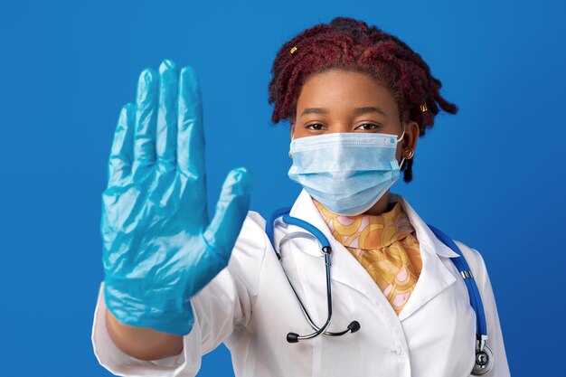 Retrato de doctora africana en bata de laboratorio con mascarilla y estetoscopio contra el fondo azul.