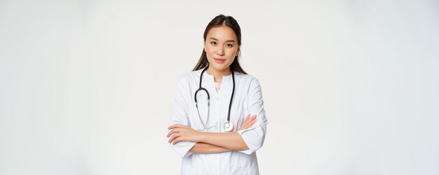 Retrato de una doctora asiática con los brazos cruzados de pie en uniforme médico y estetoscopio sonriendo a la leva