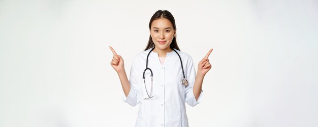 Retrato de una doctora asiática joven trabajadora de la salud en bata médica apuntando hacia los lados mostrando tw