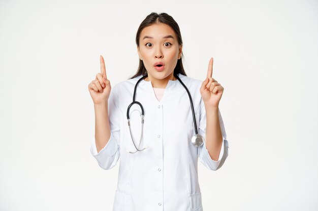 Retrato de la doctora asiática sorprendida, médico apuntando con el dedo hacia arriba, mostrando noticias asombrosas, gran promoción, fondo blanco.