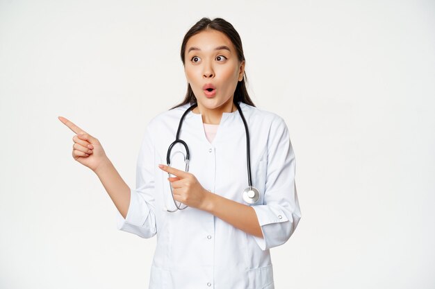 Retrato de doctora entusiasta, médico asiático apuntando y mirando a la izquierda con expresión de cara sorprendida y asombrada, de pie en bata médica contra el fondo blanco.