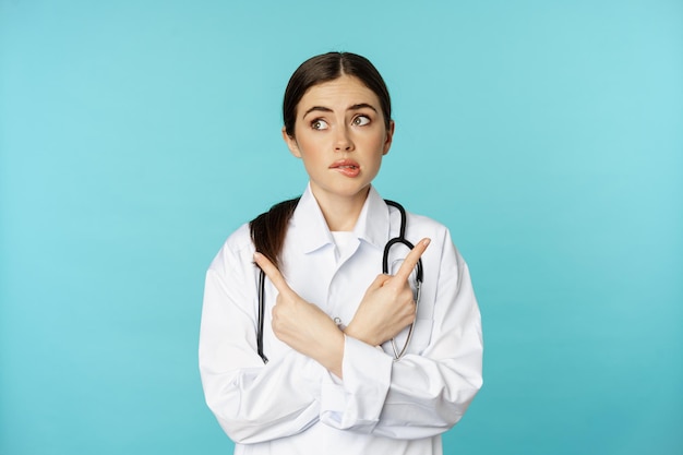 Retrato de una doctora indecisa y confundida, trabajadora de farmacia que señala de lado y se muerde el labio sin idea, no sé, parada sobre fondo azul