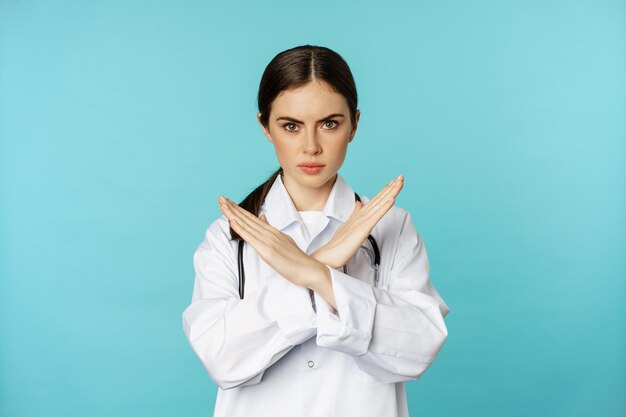 Retrato de una doctora, un médico que muestra una cruz, deja de prohibir el gesto, prohíbe algo, de pie con una bata blanca sobre un fondo turquesa