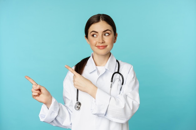 Retrato de una doctora trabajadora médica sonriente con bata blanca y estetoscopio señalando con el dedo a la izquierda...