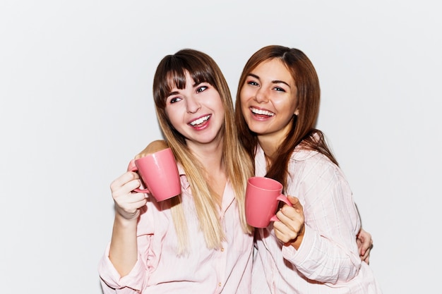 Retrato de dos alegres mujeres blancas en pijama rosa con taza de té posando de cerca. Retrato flash.