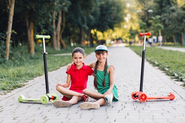 Retrato de dos amigas sentadas en la pasarela con sus patinetes en el parque