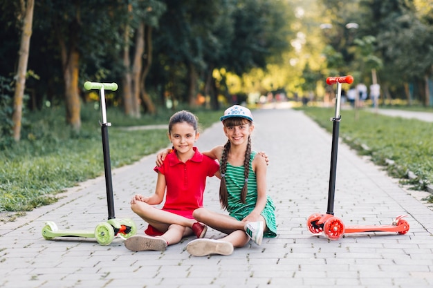 Retrato de dos amigas sentadas en la pasarela con sus patinetes en el parque