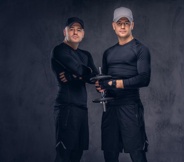 Retrato de dos apuestos jóvenes vestidos con ropa deportiva negra y gorra de béisbol sobre un fondo oscuro, sostiene una pesa.