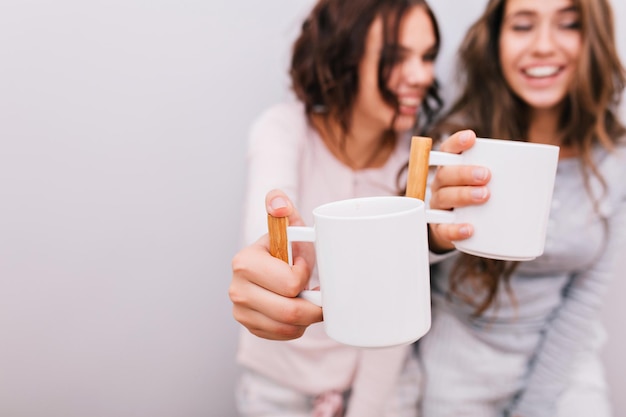 Retrato de dos chicas divertidas en pijama divirtiéndose sobre fondo de pared gris. Concéntrese en estirar las tazas blancas en sus manos.