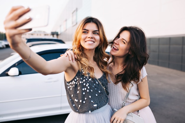 Retrato de dos chicas guapas divirtiéndose en el estacionamiento. Chica rubia haciendo selfie-retrato, morena cerca de mostrar la lengua, sonriendo.