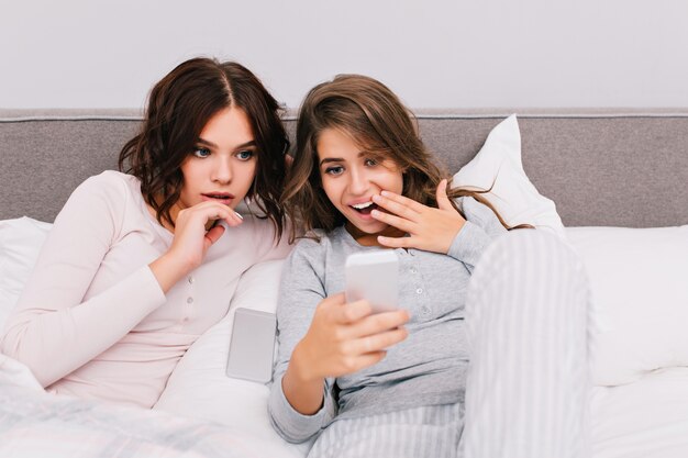 Retrato de dos chicas guapas en pijama acostado en la cama. Una niña sonriendo al teléfono, otra está asombrada.