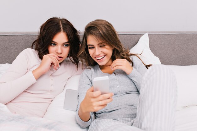 Retrato de dos chicas guapas en pijama en la cama. La chica con el pelo rizado parece asombrada, otra chica con el pelo largo sonriendo al teléfono en la mano.