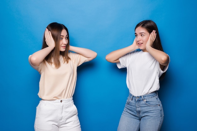 Retrato de dos divertidas mujeres de raza mixta que cubren las orejas aisladas sobre pared azul