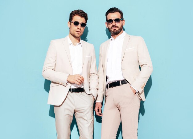 Retrato de dos guapos modelos lambersexuales con estilo inconformista confiados Hombres modernos sexy vestidos con traje elegante blanco Hombre de moda posando en el estudio cerca de la pared azul con gafas de sol