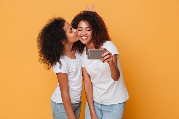 Retrato de dos hermanas afroamericanas felices tomando selfie con smartphone, lindo beso