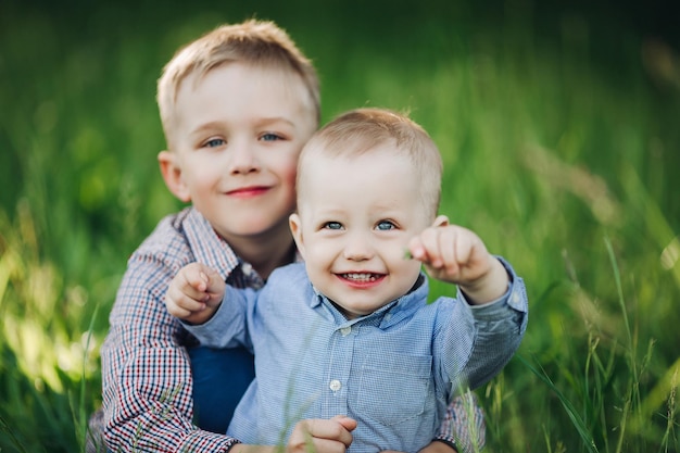 Foto gratuita retrato de dos hermanitos felices y elegantes con hermosos ojos azules jugando en el parque abrazando y mirando a la cámara niños vestidos con camisas posando sobre fondo de hierba verde