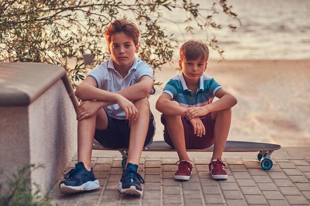 Retrato de dos hermanos pequeños sentados juntos en una patineta contra el fondo de una costa al atardecer.