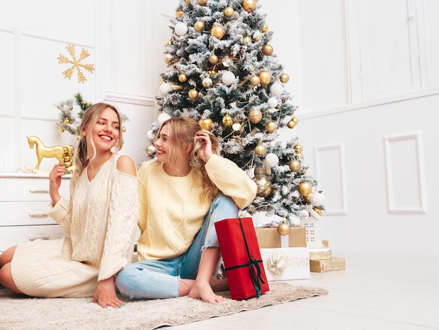 Retrato de dos hermosas mujeres rubias Modelos posando cerca del árbol de Navidad decorado en la víspera de Año Nuevo Mujer divirtiéndose lista para la celebración Amigos vestidos con cálidos suéteres de invierno