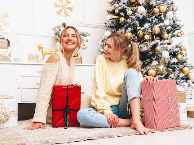 Retrato de dos hermosas mujeres rubias Modelos posando cerca del árbol de Navidad decorado en la víspera de Año Nuevo Mujer divirtiéndose lista para la celebración Amigos vestidos con cálidos suéteres de invierno