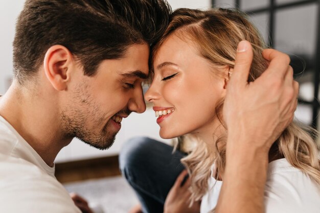 Retrato de dos hermosas personas enamoradas sonriendo con los ojos cerrados Hombre tierno tocando a su novia atractiva Chica rubia feliz en apartamentos modernos con su novio