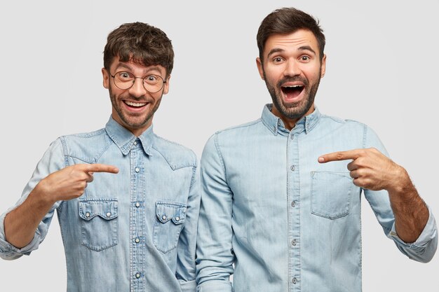 Retrato de dos hombres alegres indican con los dedos índice el uno al otro, felices de comprar nuevas chaquetas de mezclilla