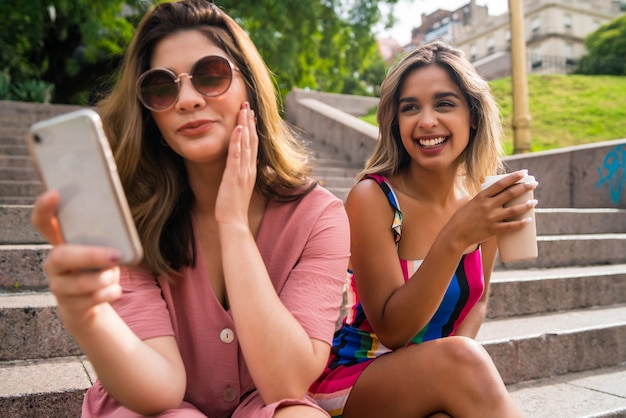 Retrato de dos jóvenes amigos disfrutando juntos y usando su teléfono móvil mientras están sentados en las escaleras al aire libre. Concepto urbano.