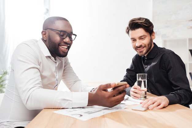 Retrato de dos jóvenes empresarios multinacionales alegres sentados en la mesa y hablando felizmente mientras trabajan juntos en la oficina aislados