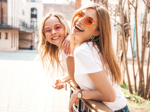 Retrato de dos jóvenes hermosas rubias sonrientes chicas hipster en ropa de moda verano camiseta blanca. . Modelos positivos divirtiéndose en gafas de sol.