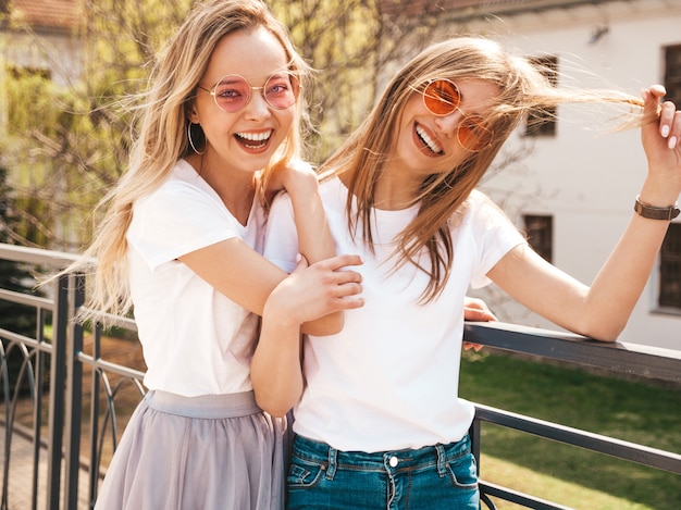 Foto gratuita retrato de dos jóvenes hermosas rubias sonrientes chicas hipster en ropa de moda verano camiseta blanca. . modelos positivos divirtiéndose en gafas de sol.