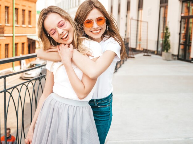 Retrato de dos jóvenes hermosas rubias sonrientes chicas hipster en ropa de moda verano camiseta blanca. . Modelos positivos divirtiéndose en gafas de sol.