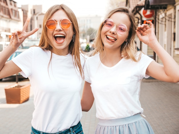 Retrato de dos jóvenes hermosas rubias sonrientes chicas hipster en ropa de moda verano camiseta blanca. . Modelos positivos divirtiéndose Muestra signo de paz