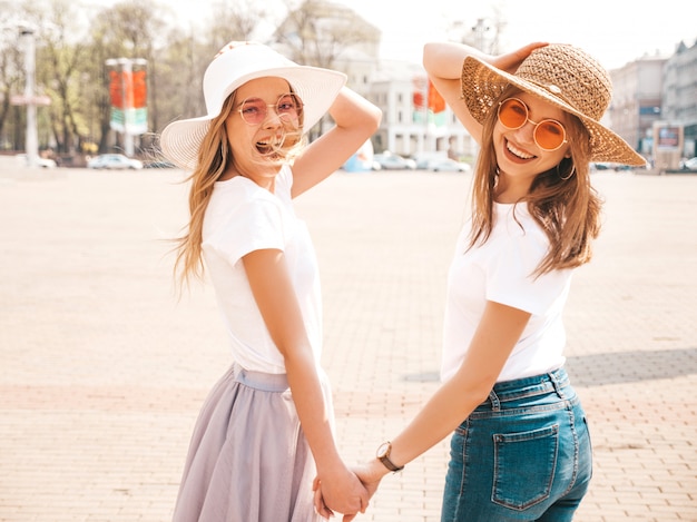 Retrato de dos jóvenes hermosas rubias sonrientes chicas hipster en ropa de moda verano camiseta blanca. . Modelos positivos tomados de la mano