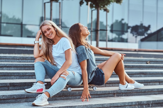 Retrato de dos jóvenes hipsters sentadas juntas en una patineta en pasos sobre un fondo del rascacielos.